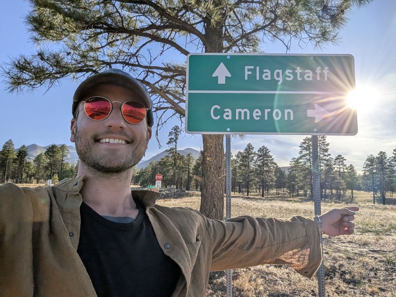Cameron smiling beside a green road sign pointing toward Flagstaff and Cameron in the Arizona high country