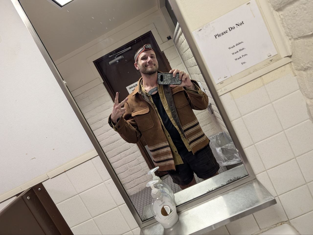 Cameron taking a mirror selfie in a diner bathroom wearing a brown jacket and headlamp