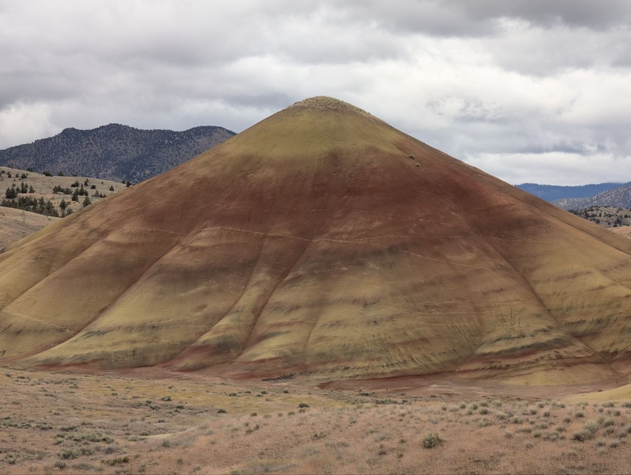 A conical Painted Hills formation with red and yellow bands under cloudy sky