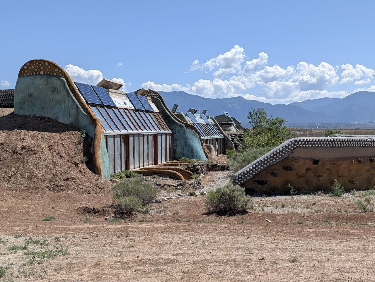 A row of Earthship buildings with solar panels and greenhouse glass facing the mountains